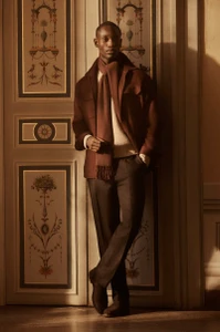 Charles Oduro posing in a brown jacket, matching knit scarf, cream sweater, and dark trousers against an ornate painted door.