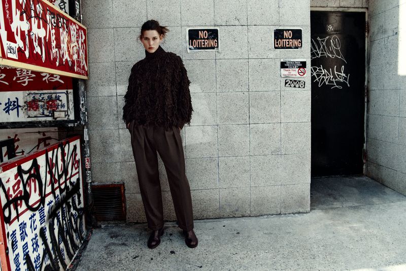 Fashionable woman in textured top and trousers posing against an urban wall with graffiti and signs in a street scene.