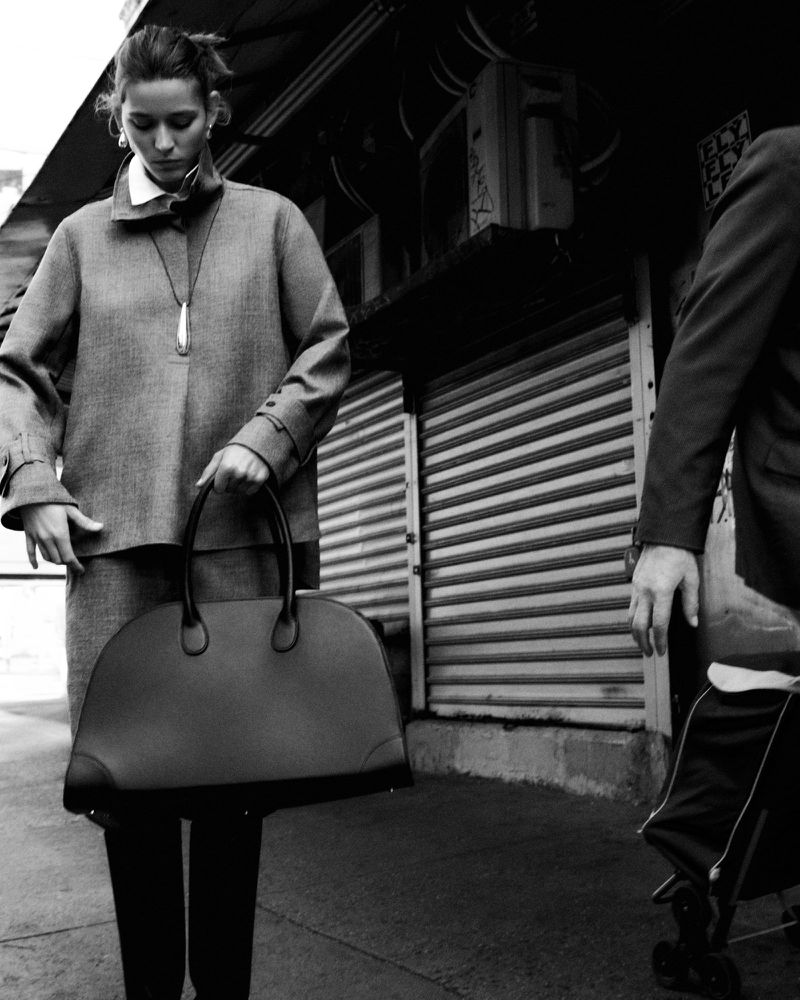 A woman in a monochrome urban setting carries a large handbag with a man partially visible in the background, creating a stylish, understated scene.