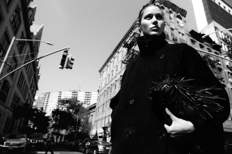 Black and white fashion shot of a woman in a dark coat holding a textured bag on a city street with buildings and traffic signals.