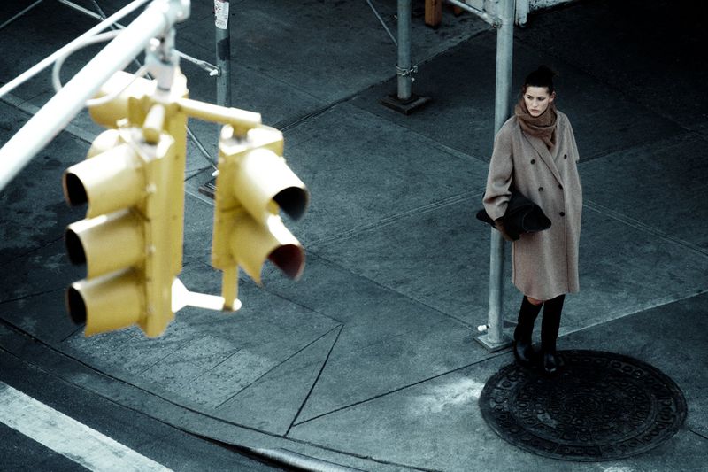 Fashionable woman in beige coat stands at city street corner by yellow traffic light, holding a black hat and purse, urban street scene.