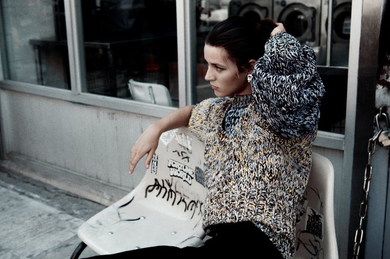 A woman with her hair up, wearing a knit sweater and sitting on a graffiti-covered chair in front of a laundromat, looking off to the side.