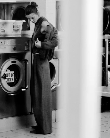 Black and white shot of a woman in a laundromat, adjusting her sweater, standing in front of a row of washing machines.
