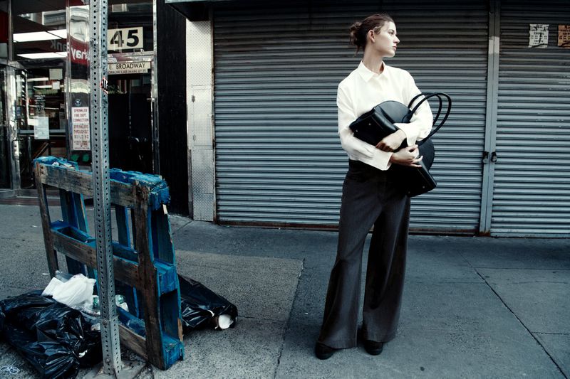 Fashionable woman stands on a city sidewalk wearing a white blouse and grey slacks, carrying a black bag in front of a roll-up door.