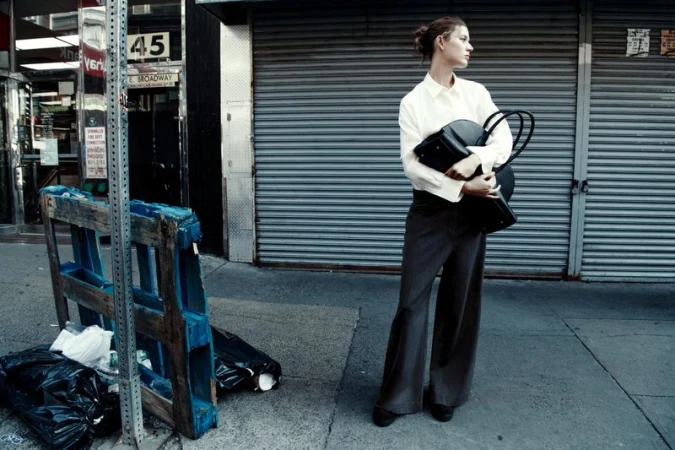 Fashionable woman stands on a city sidewalk wearing a white blouse and grey slacks, carrying a black bag in front of a roll-up door.