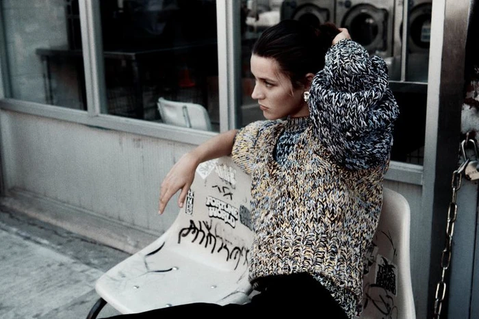 Model Vivienne Rohner adjusting hair while seated in a patterned knit sweater outside a storefront for COS Pre-Winter 2024