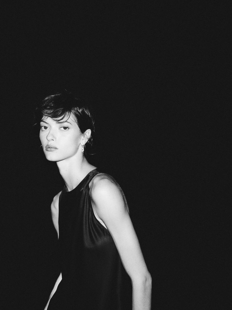 Monochromatic portrait of a young woman with short hair in a sleeveless dress, posing against a dark background in a studio setting.