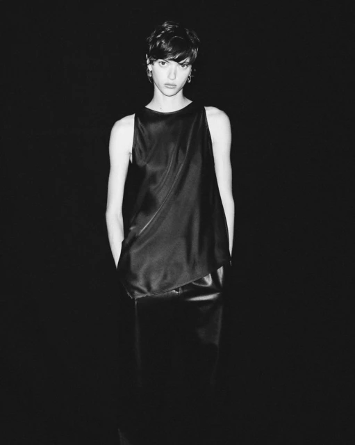 Monochromatic portrait of an adult female with short brown hair wearing a black satin top and skirt against a black background.