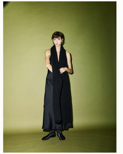 A woman in a full-length black dress with a long knitted black scarf poses in front of an olive-green backdrop in a studio setting.