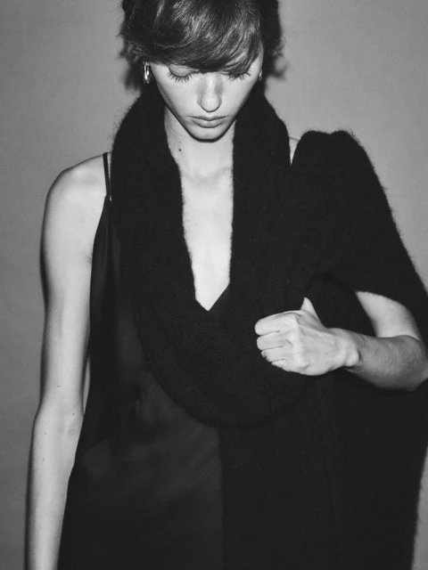 Monochrome portrait of a woman with short hair looking down, wearing a black dress and a large black scarf draped around her neck.
