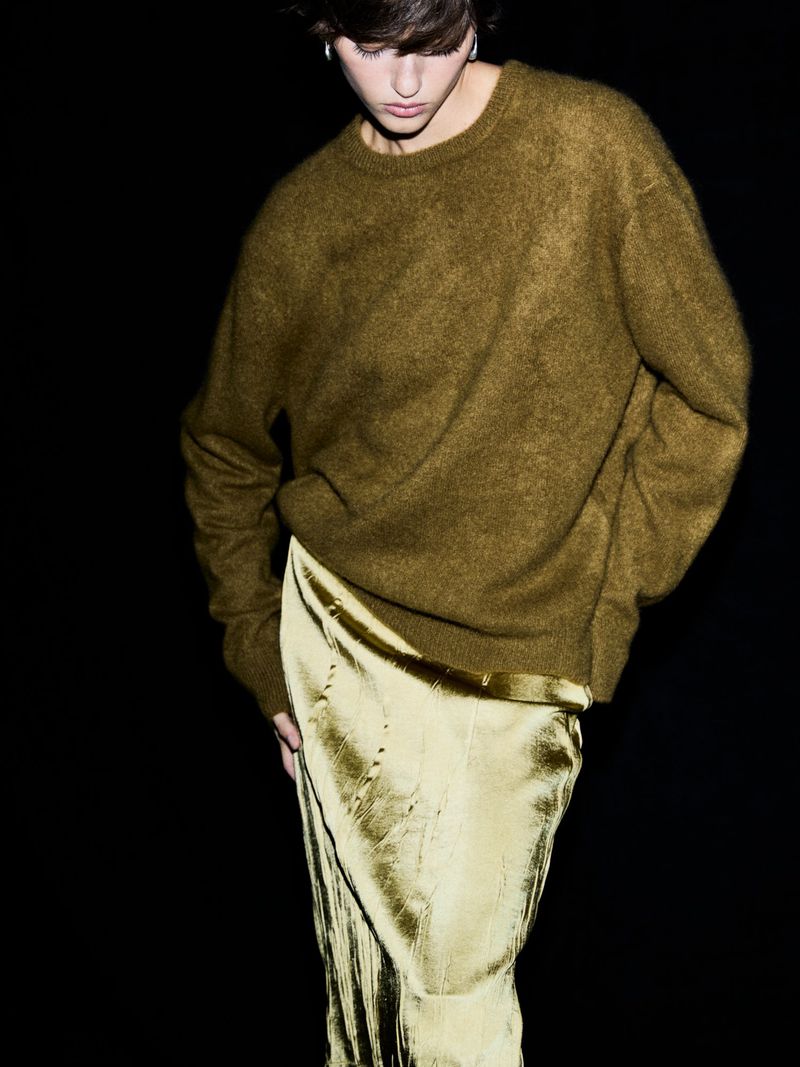 Person wearing an olive green sweater and a shimmering gold skirt against a dark backdrop in a studio fashion shot.