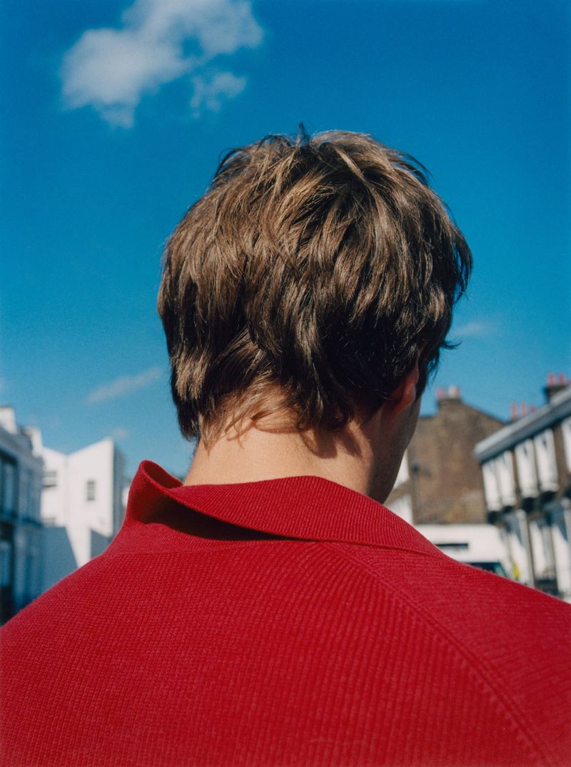 From behind, an adult male with short brown hair wears a red polo shirt against a bright blue sky and urban buildings.
