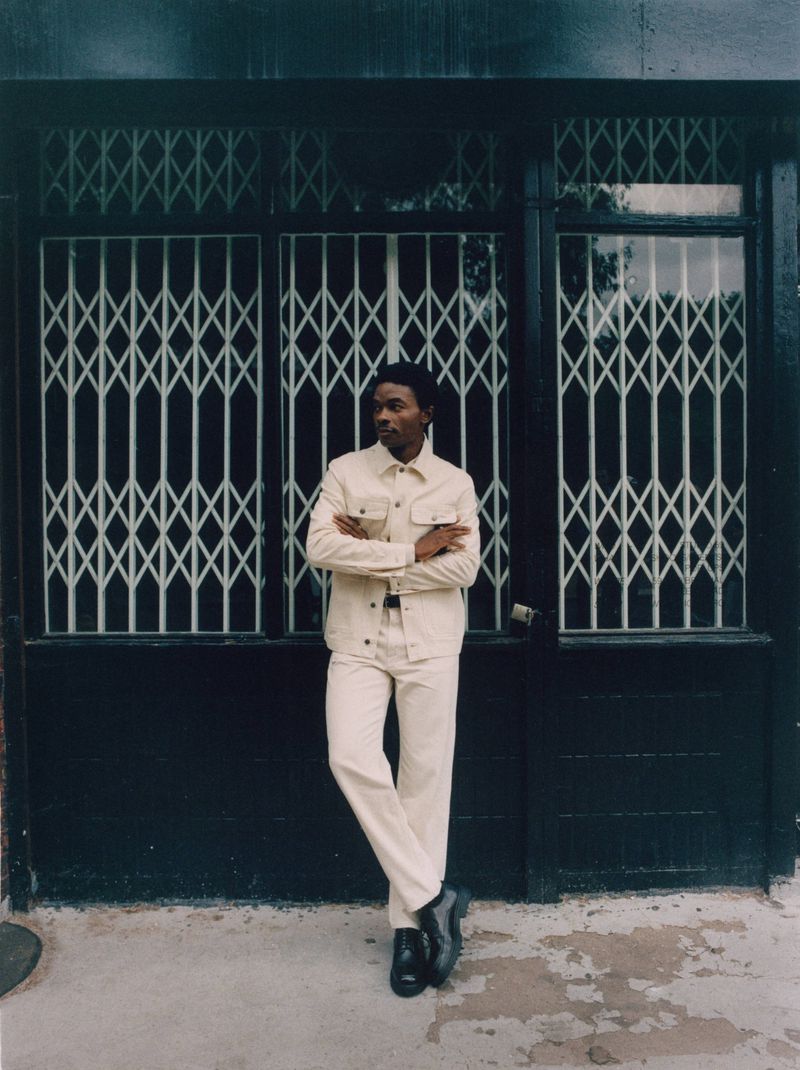A slender adult Black man poses outdoors in a cream-colored denim jacket and matching jeans, leaning against a black metal security gate.