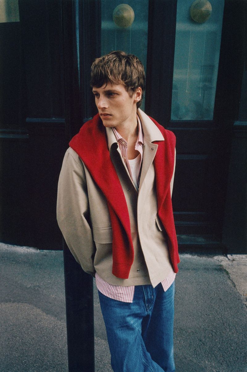 A young adult man in a beige jacket and blue jeans leans against a dark post outdoors, with a red sweater draped over his shoulders.