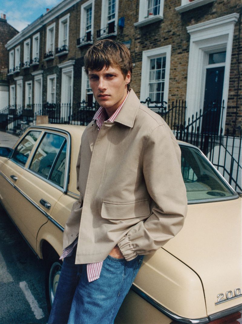 An adult male fashion model leans against a classic car in an urban environment, wearing a tan jacket and jeans.