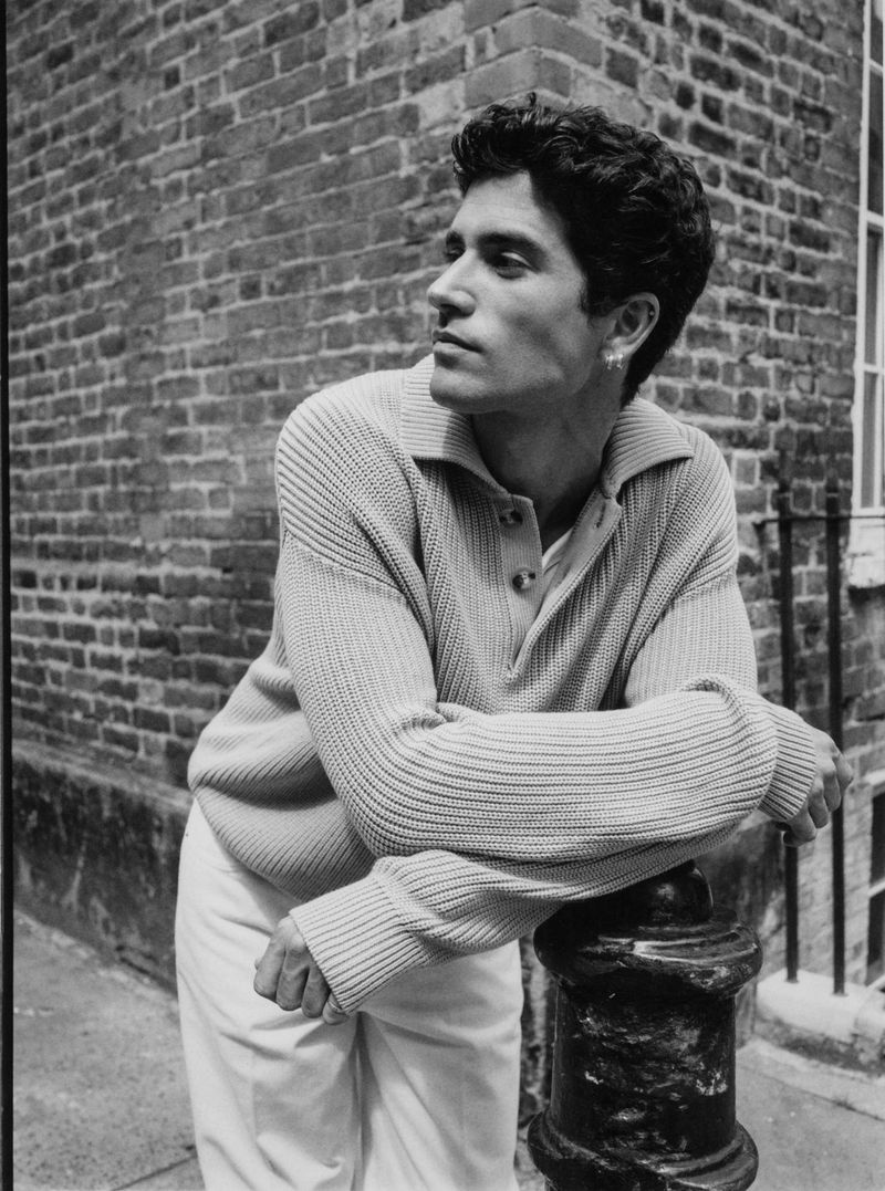 Black and white portrait of a man leaning on a bollard with a brick wall background, wearing a collared button-down sweater and light pants.