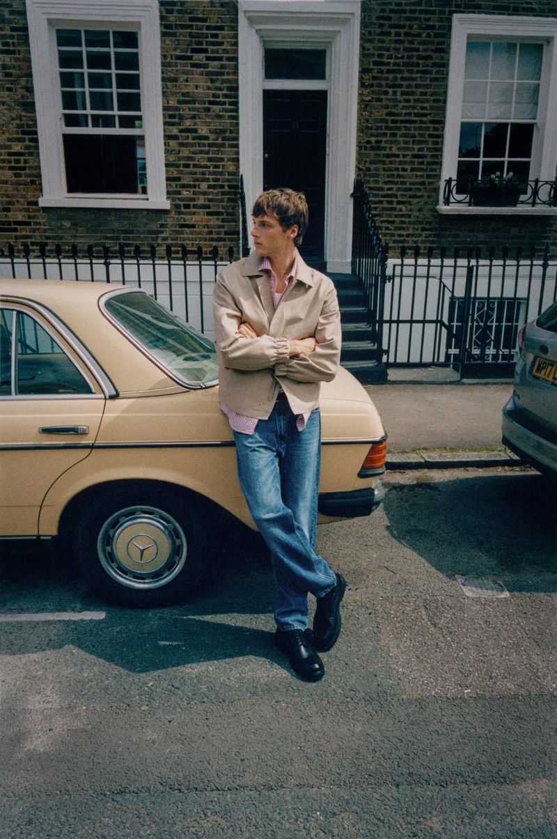 Young adult male stands beside a tan vintage car on a sunny street with brick buildings in background, wearing casual attire.