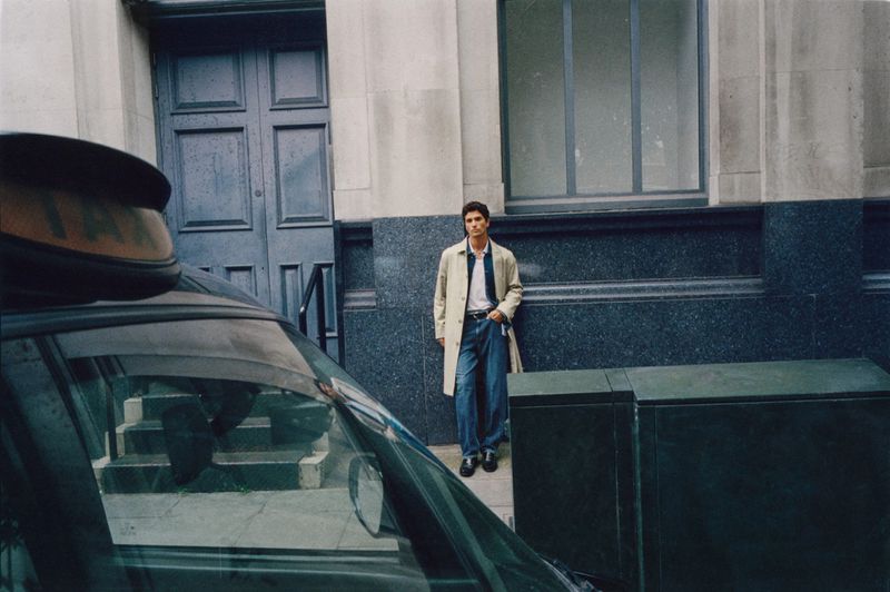 A man in a trench coat stands near a building while a taxi's windshield is visible in the foreground, creating an urban street scene.