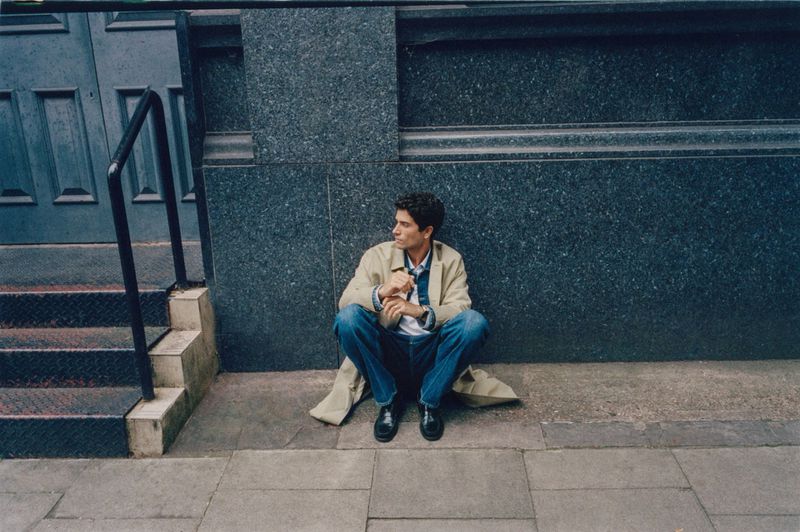 Man in beige coat and jeans sits on city sidewalk next to stairs and dark blue wall, looking pensive in outdoor daylight setting.