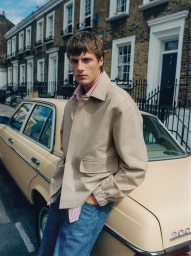 Model Hugh Laughton-Scott leans against a cream vintage car wearing a beige jacket and blue denim jeans on a residential street.