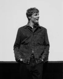 Monochrome portrait of a man wearing a dark denim jacket with hands in pockets against a plain white wall