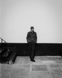A man wearing a denim jacket and jeans stands leaning against a white wall next to a staircase.