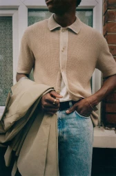 Close-up of a Black man in a beige knit polo shirt and light wash denim jeans holding a folded trench coat outside a brick building.