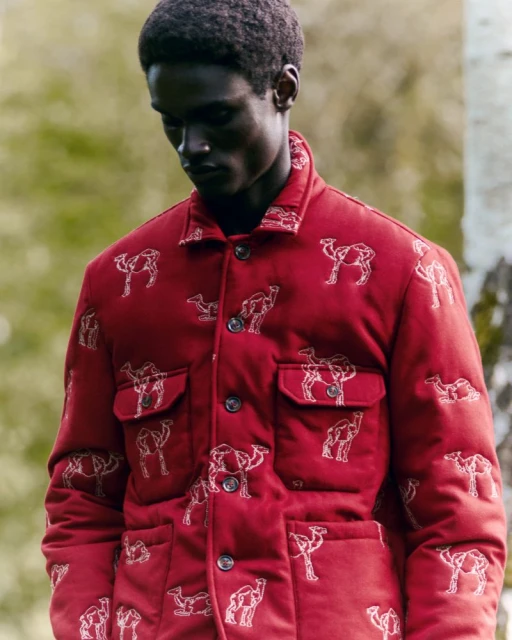 A man in a red patterned jacket stands in a grassy field. The jacket has buttons and pockets. Trees are visible in the background.