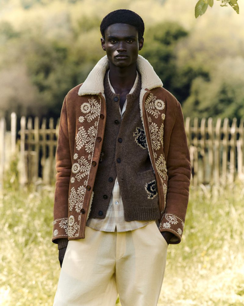 A dark-skinned man stands outside wearing a brown jacket, buttoned brown cardigan, cream pants, and a black cap in a medium shot.