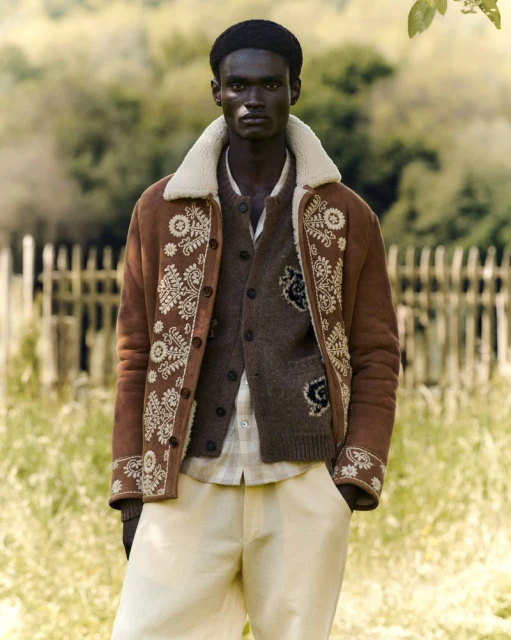 A dark-skinned man stands outside wearing a brown jacket, buttoned brown cardigan, cream pants, and a black cap in a medium shot.