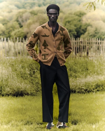 A brown-skinned adult male model wears a tan cardigan and dark pants in a grassy outdoor setting with a blurred forest background.