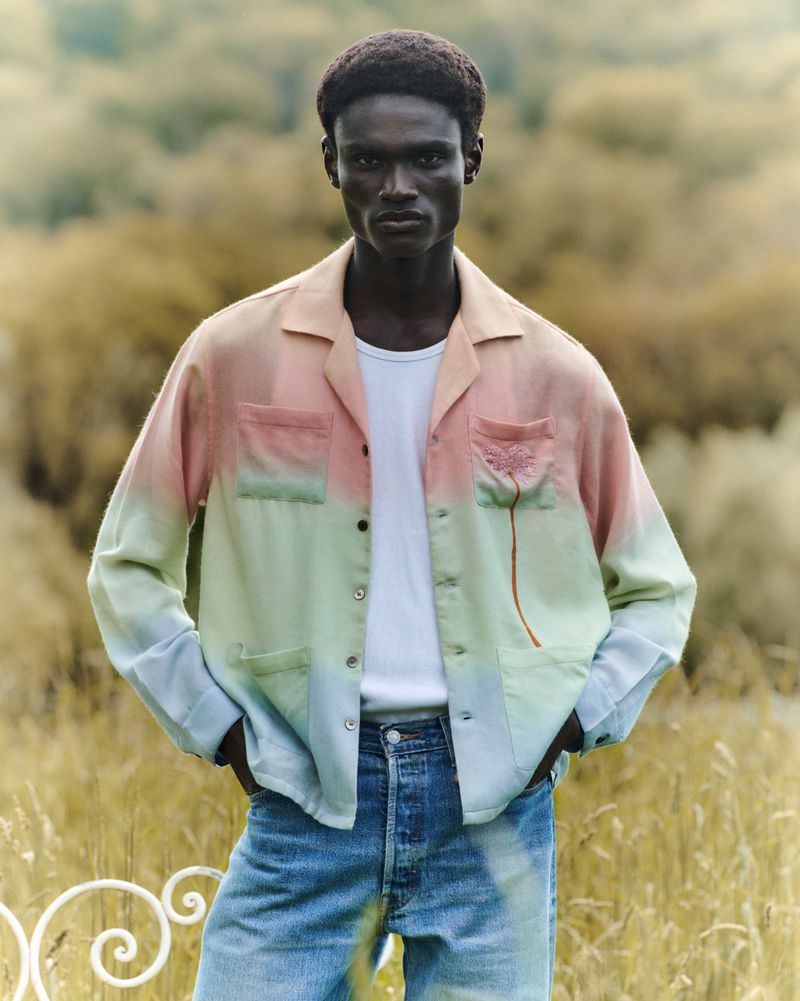 An adult male model poses outdoors, wearing an open ombre button-down shirt, a white t-shirt, and blue jeans. Tall grass is in the background.