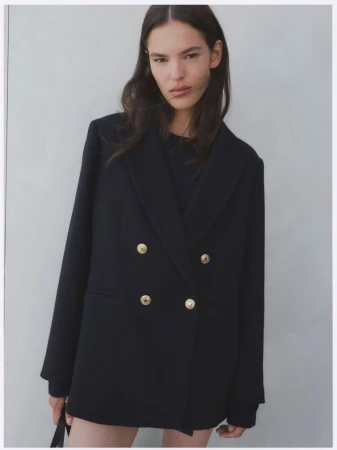 Model wearing an oversized black double-breasted blazer with gold buttons against a neutral grey studio background.