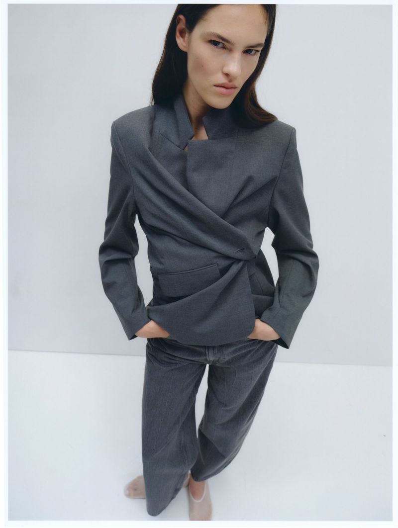Woman poses in a studio, wearing a grey jacket and wide-legged pants. The jacket is draped across the front. Studio lighting.