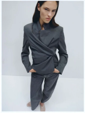 Woman poses in a studio, wearing a grey jacket and wide-legged pants. The jacket is draped across the front. Studio lighting.