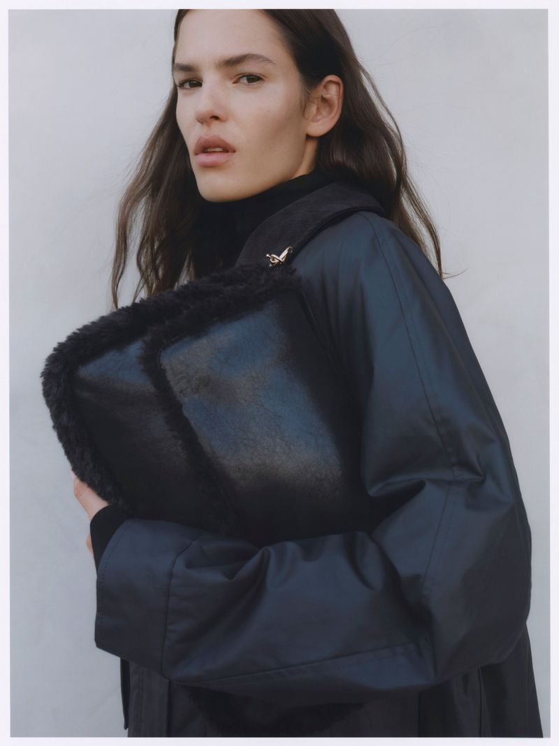 A woman in a navy coat holds a black fur-trimmed bag in front of a plain background, showcasing fashion and style.
