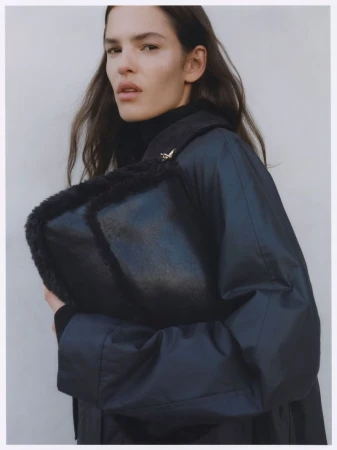 A woman in a navy coat holds a black fur-trimmed bag in front of a plain background, showcasing fashion and style.