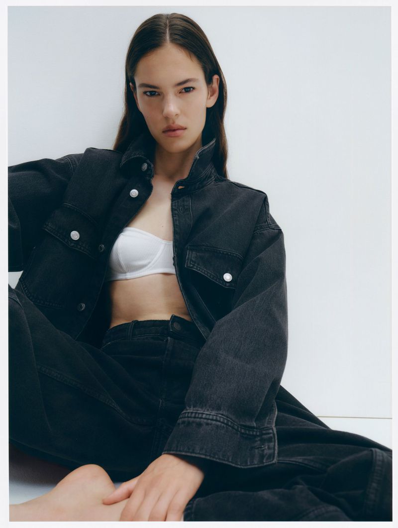 Adult female in dark denim jacket and pants, seated against a white wall, wearing a white bra, shot in soft studio lighting.