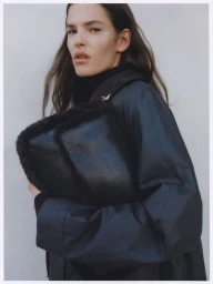 Woman wearing a black coat holding a large textured black bag against a light grey background