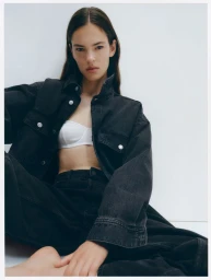 Model seated wearing a black denim jacket and matching trousers over a white top against a white studio background.