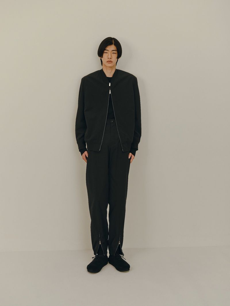 Full-length shot of an Asian man in a black bomber jacket and black trousers posing against an off-white studio backdrop for fashion.