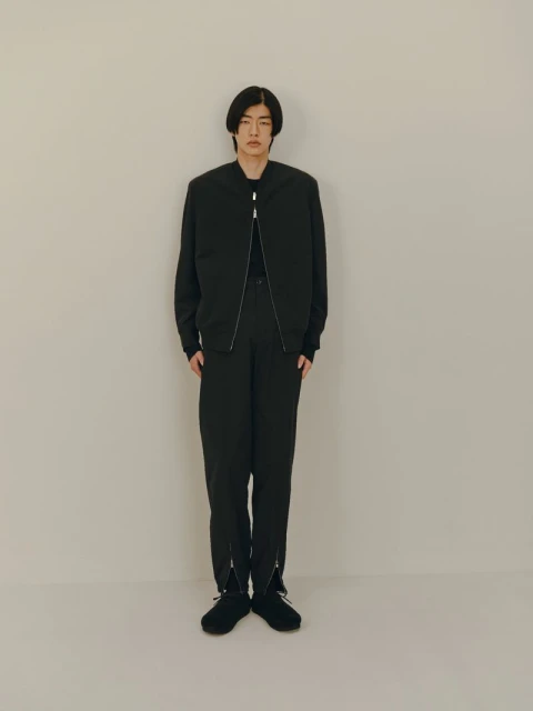 Full-length shot of an Asian man in a black bomber jacket and black trousers posing against an off-white studio backdrop for fashion.