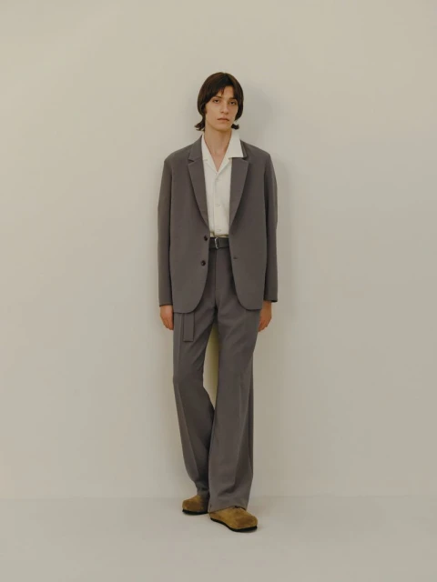 Full-length shot of an adult male model in a grey suit and tan shoes posing against a light beige studio backdrop with soft, neutral lighting.