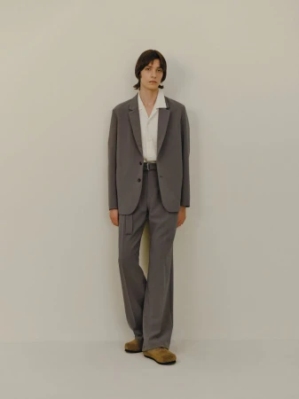 Full-length shot of an adult male model in a grey suit and tan shoes posing against a light beige studio backdrop with soft, neutral lighting.
