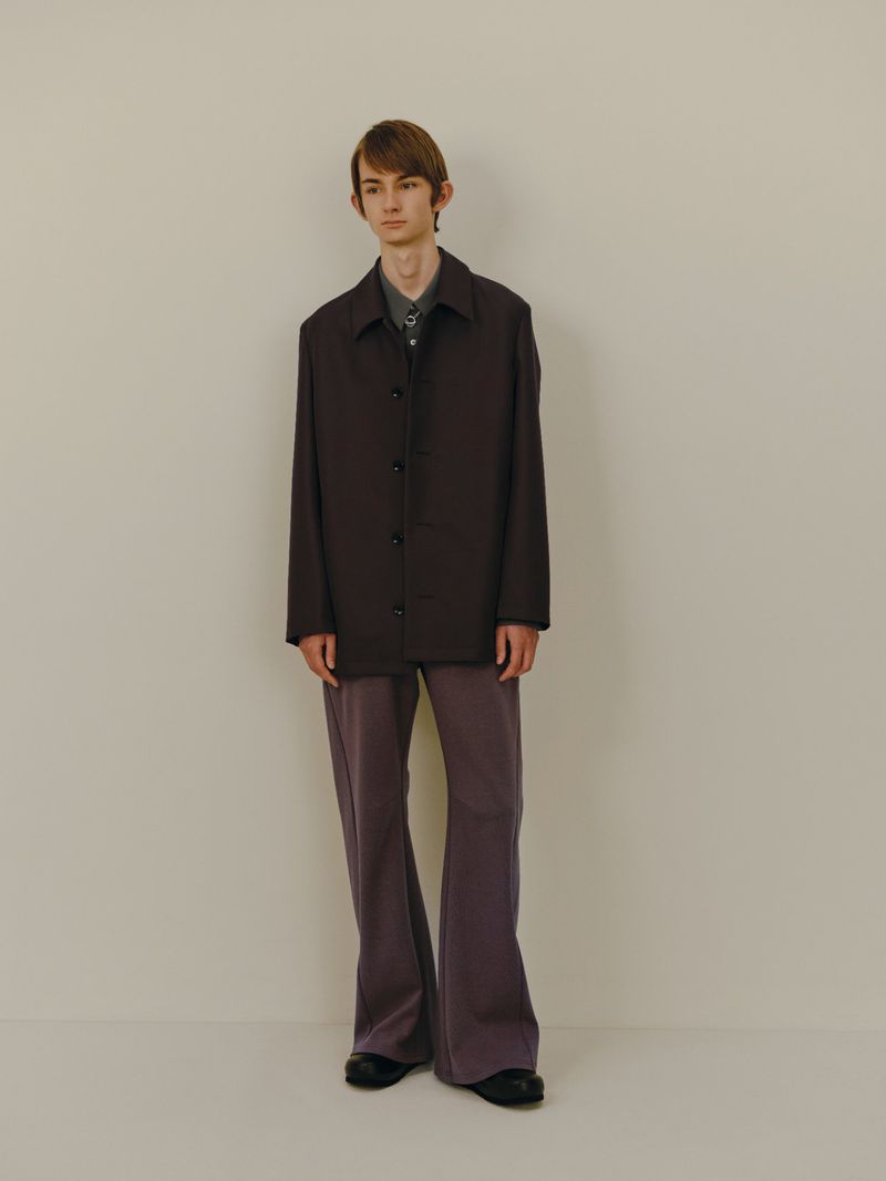 A slender man in a brown button-down jacket, a gray collared shirt, and flared pants stands against an off-white backdrop in a studio shot.