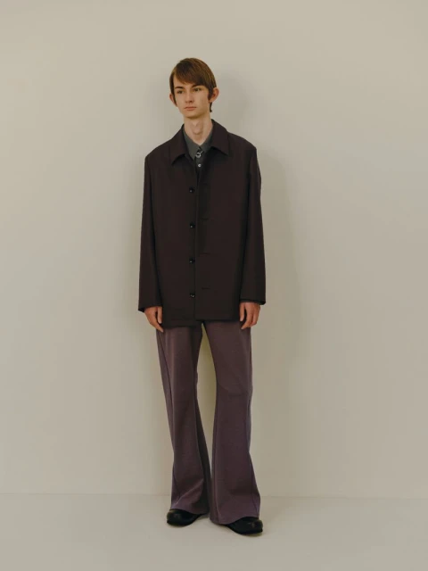 A slender man in a brown button-down jacket, a gray collared shirt, and flared pants stands against an off-white backdrop in a studio shot.