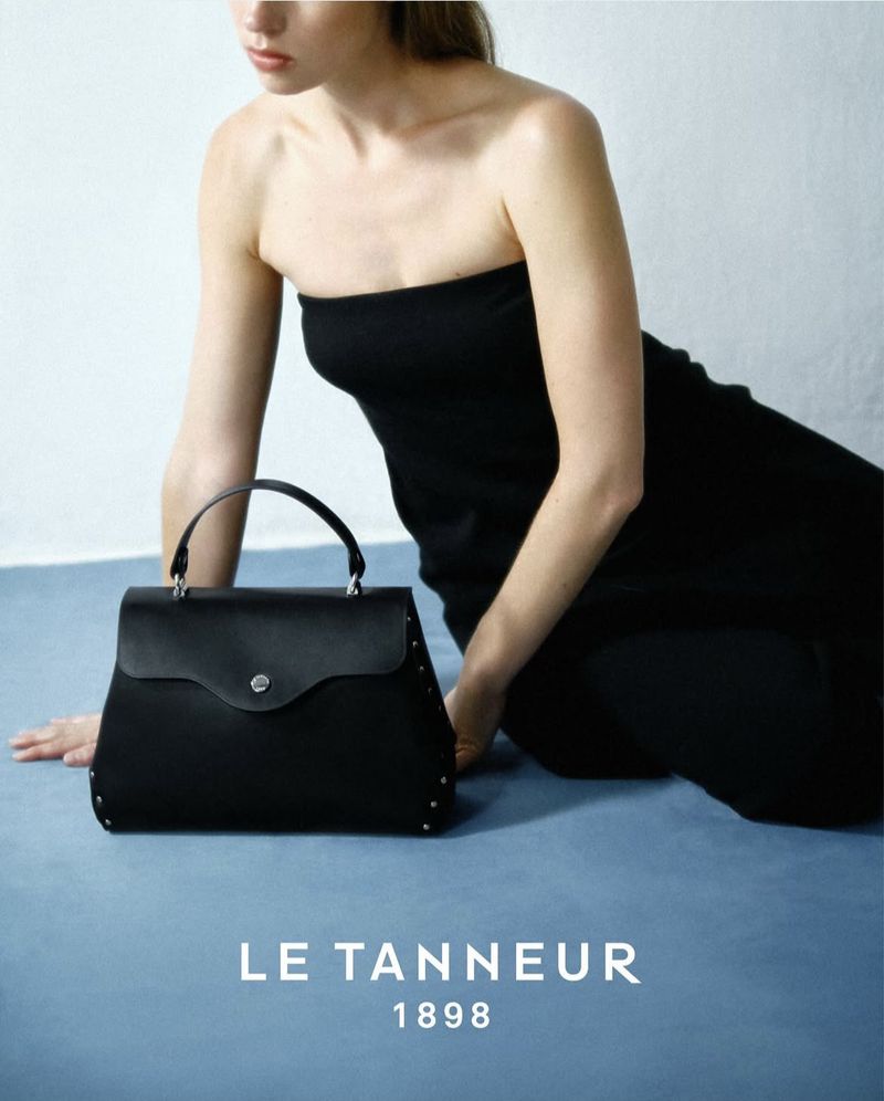 Woman poses with black leather handbag in a studio shot. The handbag features a top handle and studded details on a light blue backdrop.