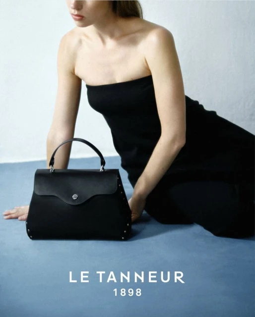 Woman poses with black leather handbag in a studio shot. The handbag features a top handle and studded details on a light blue backdrop.