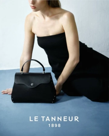 Woman poses with black leather handbag in a studio shot. The handbag features a top handle and studded details on a light blue backdrop.