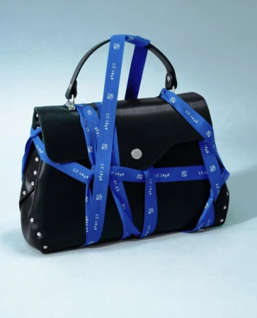 Black leather top-handle handbag wrapped crisscross in bright blue branded ribbon against a light blue studio background.